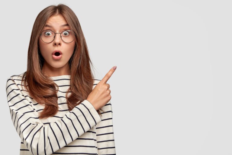 Horizontal shot of beautiful young Caucasian female with surprised expression, points at upper right corner, dressed in striped sweater, wears round spectacles, isolated over white background