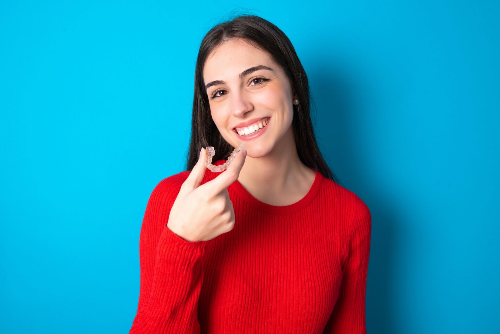 young brunette girl wearing red T-shirt against blue wall holding an invisible aligner ready to use it. Dental healthcare and confidence concept.